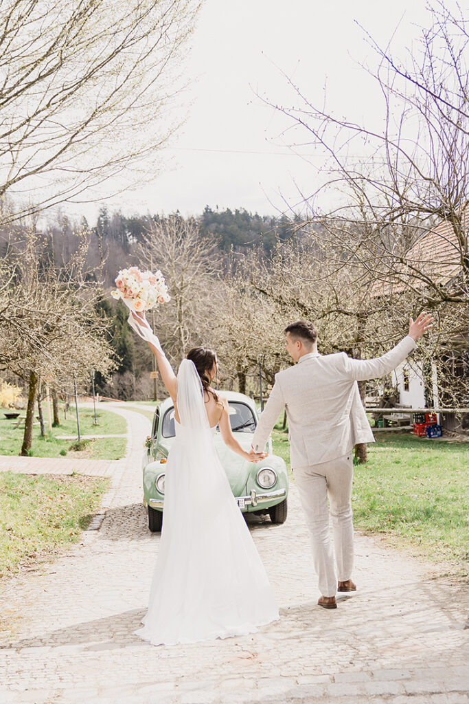 Das Brautpaar geht Hand in Hand auf einer Allee, der Oldtimer steht im Hintergrund.