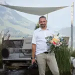 Hochzeitsfotograf Niederbayern – Kosta Mücke mit Kamera und Blumenstrauß vor alpiner Kulisse