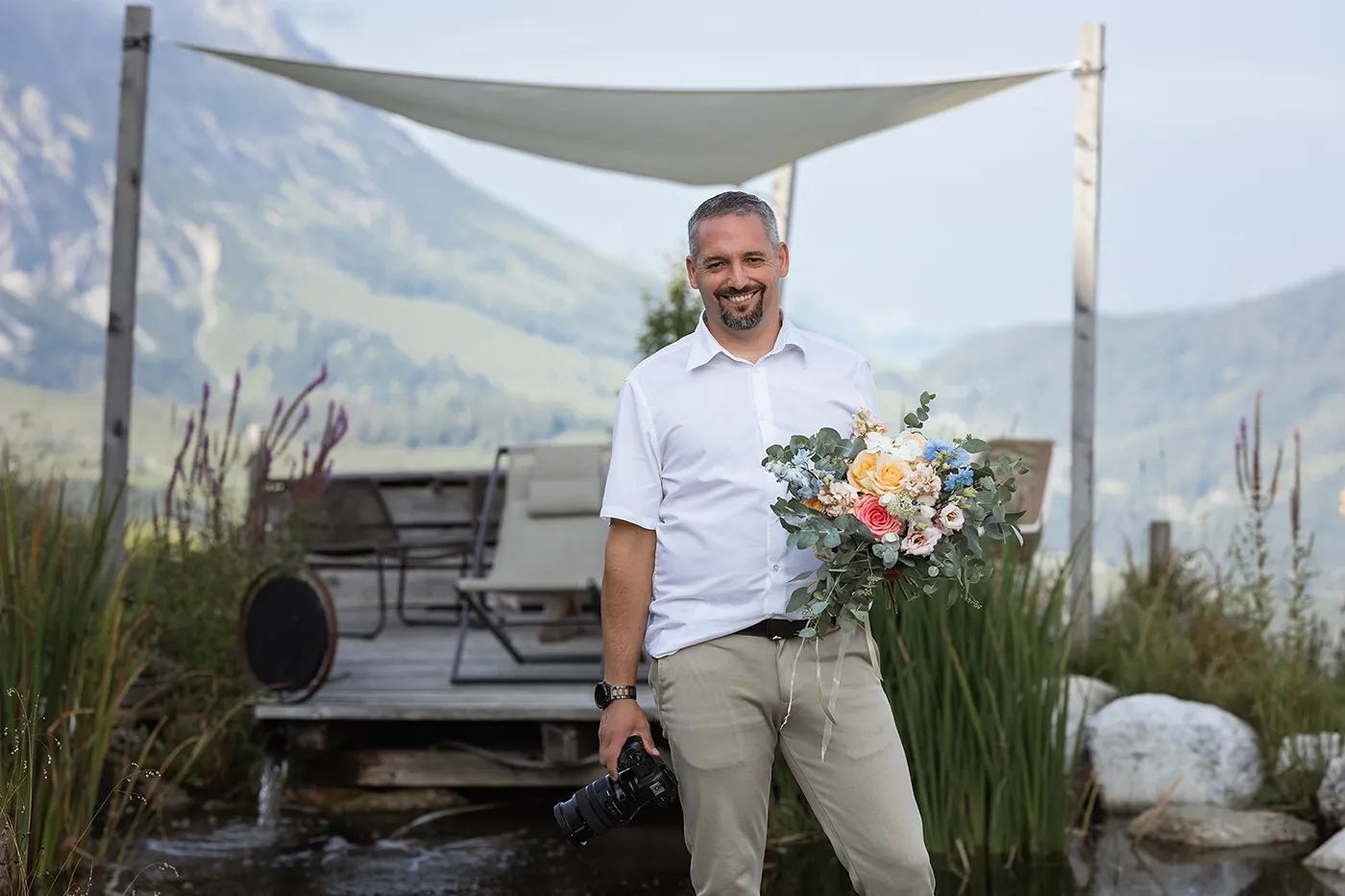 Hochzeitsfotograf Niederbayern – Kosta Mücke mit Kamera und Blumenstrauß vor alpiner Kulisse