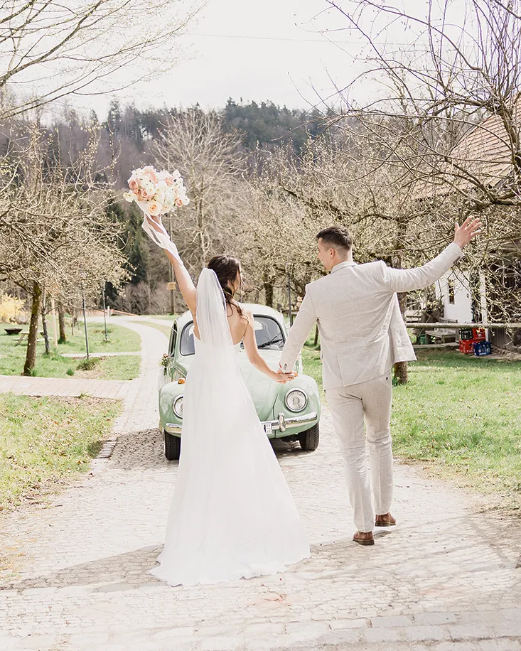 Brautpaar läuft Hand in Hand über den Weg am Grandsberger Hof, mit Blumenstrauß und grünem Oldtimer im Hintergrund.
