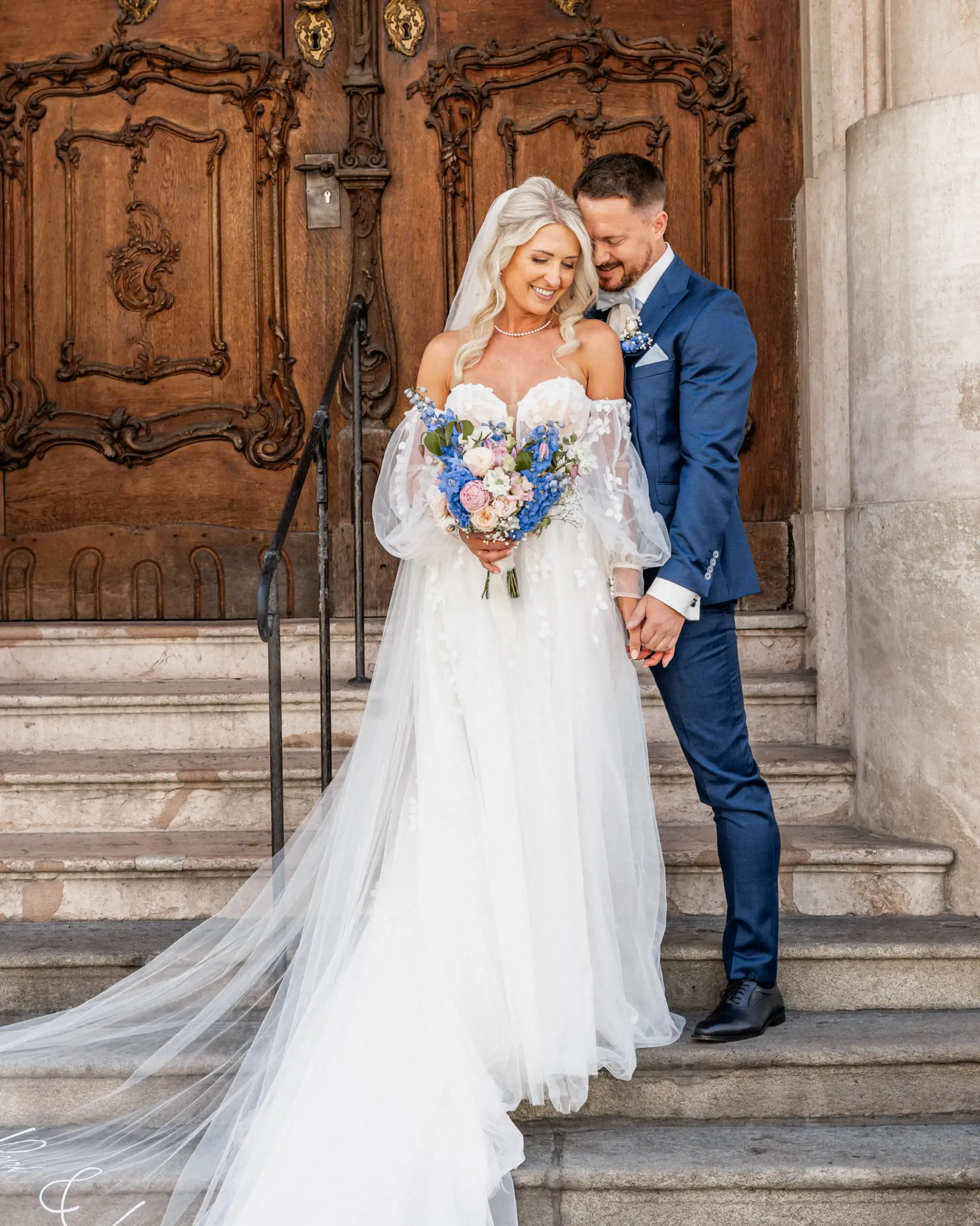 Brautpaar steht lächelnd auf den Treppen vor einer alten Holztür in Passau – ein Moment voller Liebe und Eleganz.