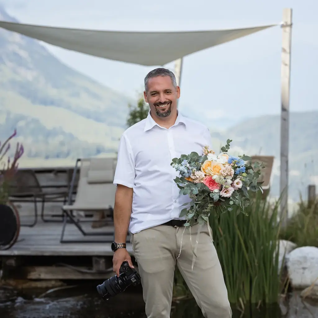 Kosta Mücke, Hochzeitsfotograf aus Passau, bei einem Portrait auf der Tiergartenalm mit Kamera und Blumenstrauß. 