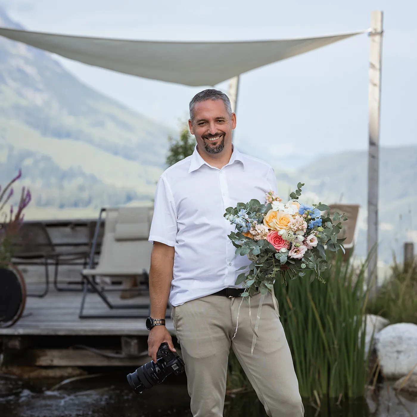 Kosta Mücke, Hochzeitsfotograf aus Passau, bei einem Portrait auf der Tiergartenalm mit Kamera und Blumenstrauß.