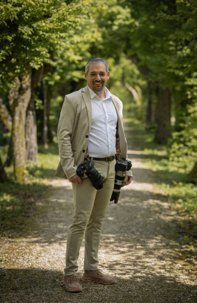 Kosta Mücke, Hochzeitsfotograf aus Passau, lächelnd mit zwei Kameras auf einem Weg zwischen Bäumen
